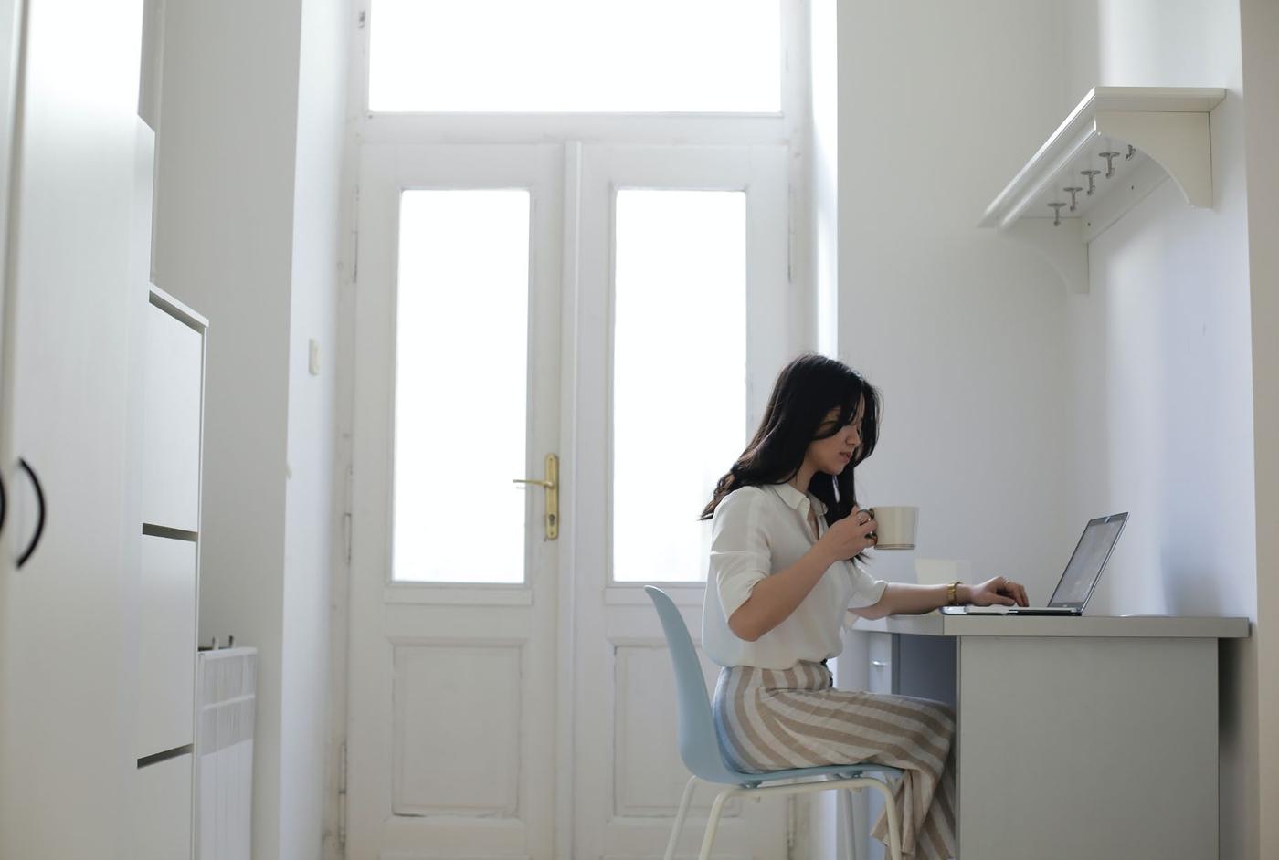 Mulher trabalha em casa com um notebook, sentada a uma mesa clara ao lado de portas brancas.